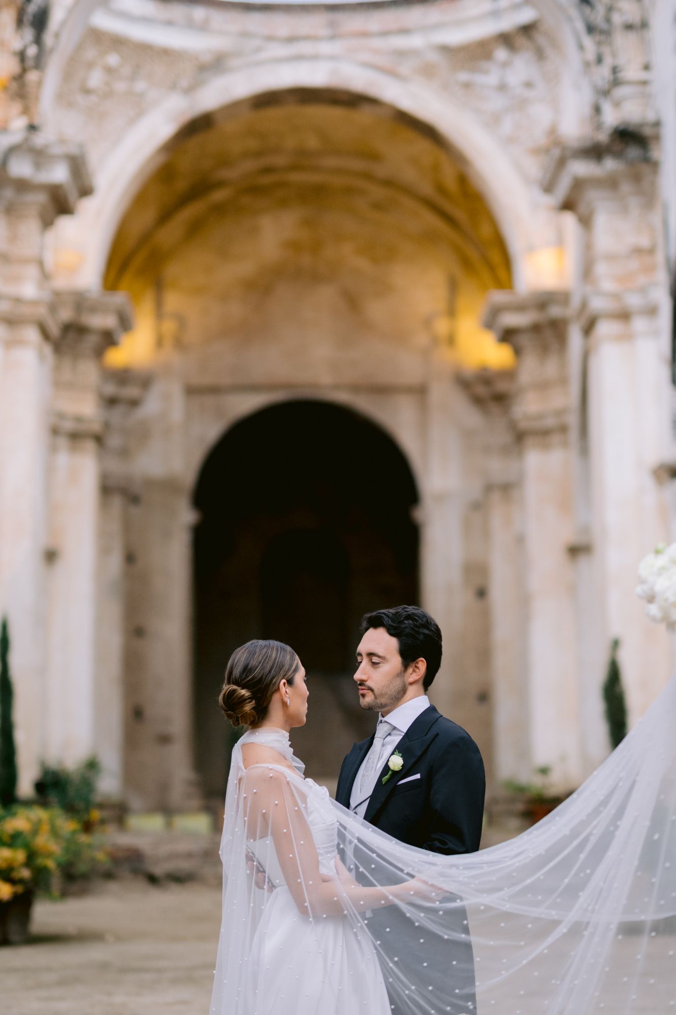 Romantic newlyweds embrace under historic stone archway with flowing veil details.
