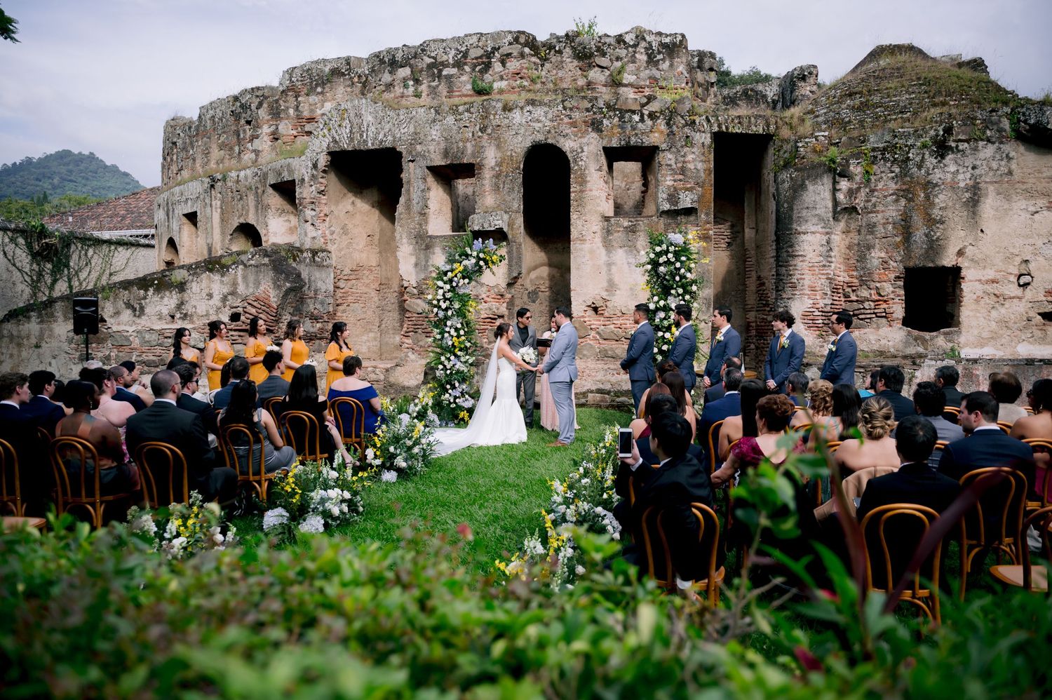 Wedding ceremony at historic ruins site with decorated arches and seated guests.
