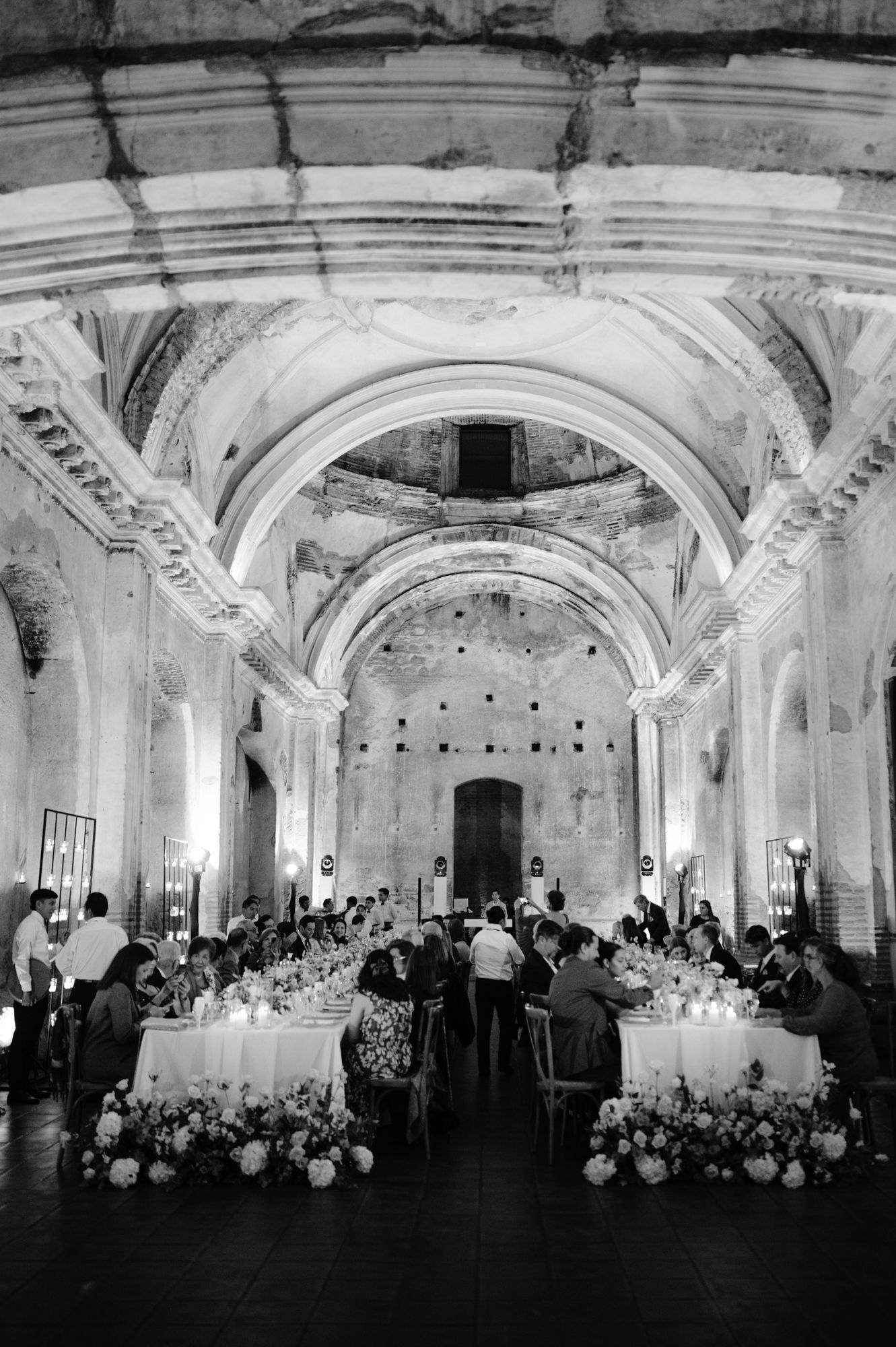Black and white photo of wedding reception in grand vaulted ceiling venue with long tables.
