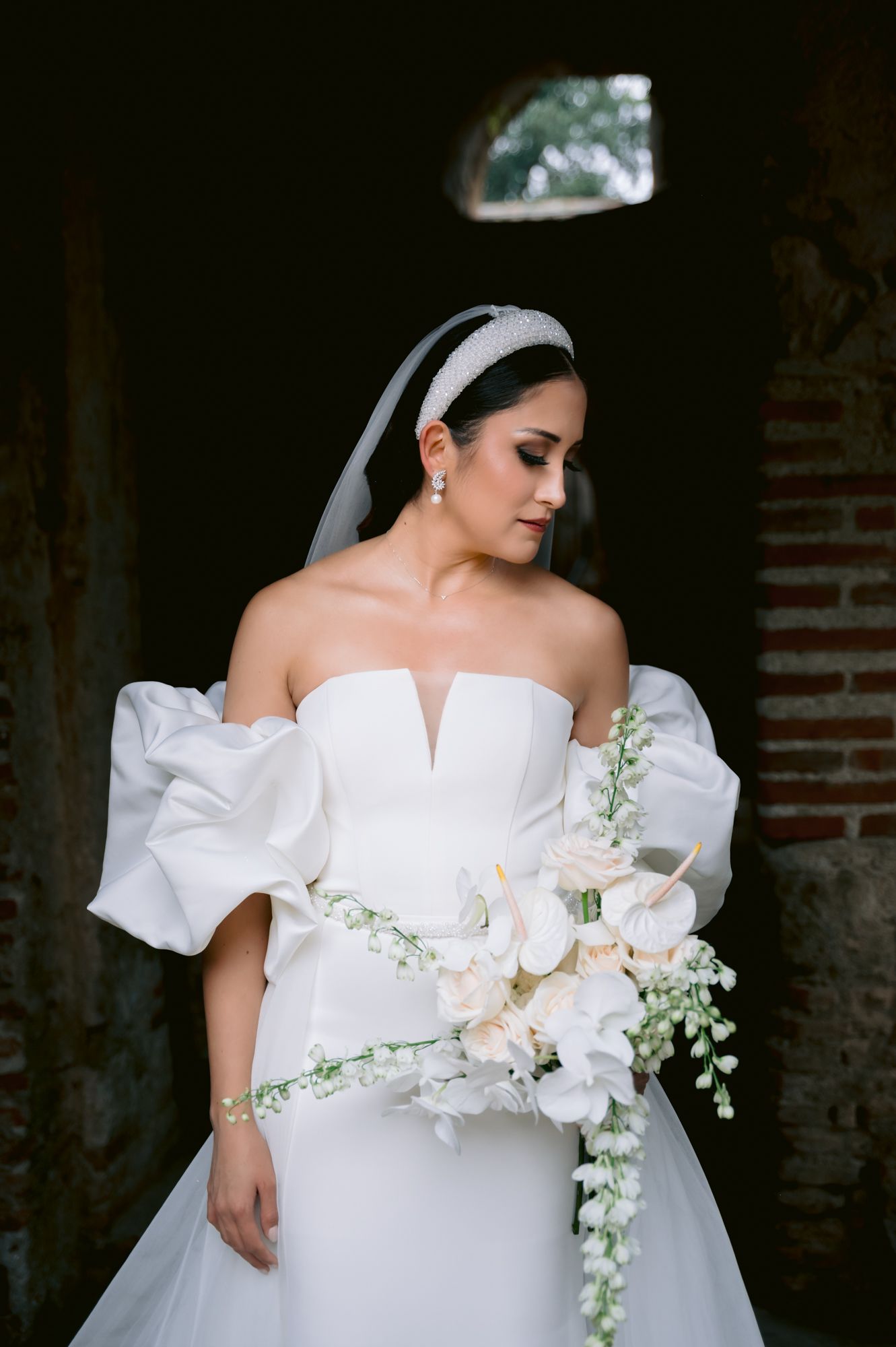 Bride in structured wedding gown with dramatic sleeves holds cascading white floral bouquet.