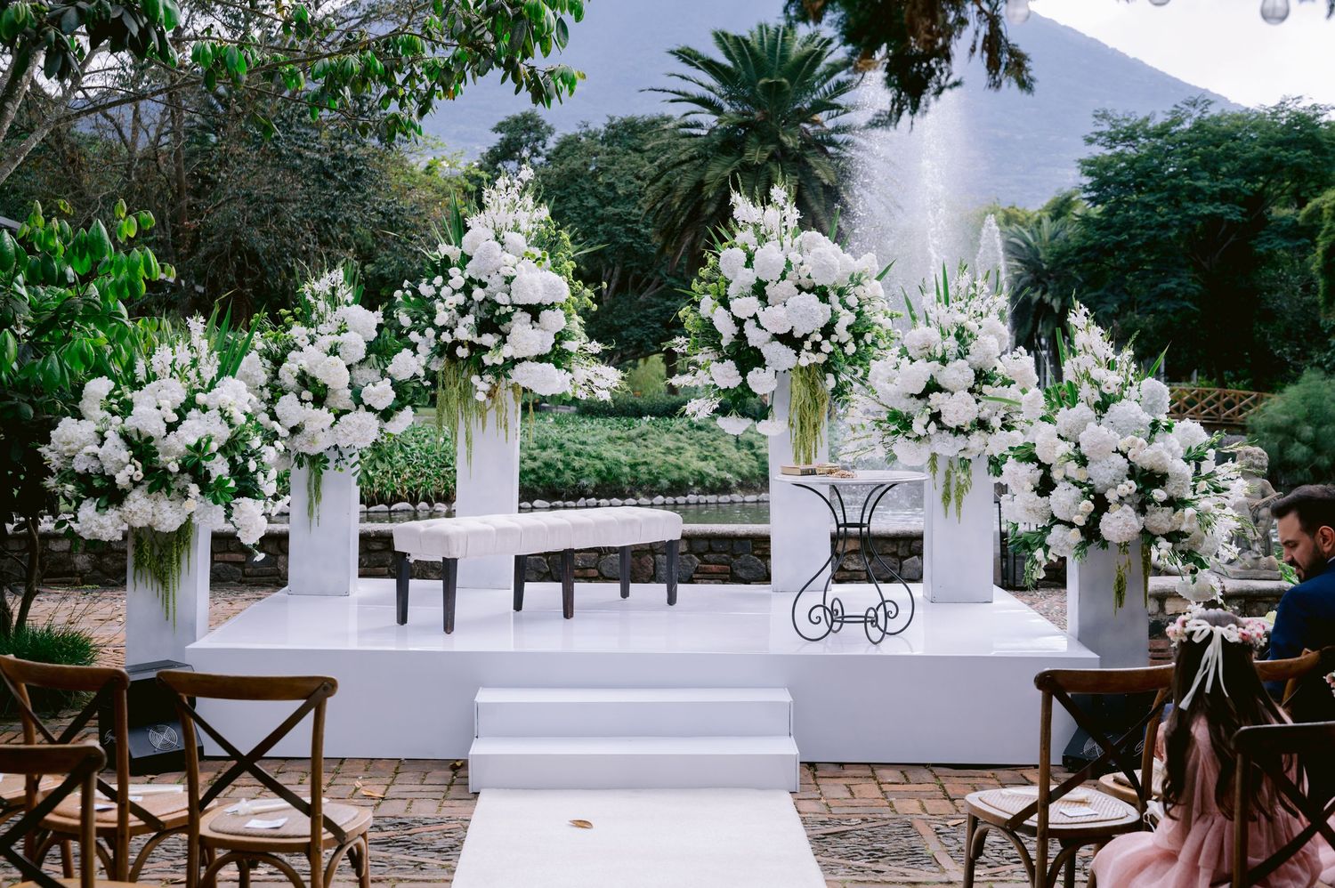 Elegant outdoor wedding ceremony setup with white floral arrangements on pedestals and wooden chairs.