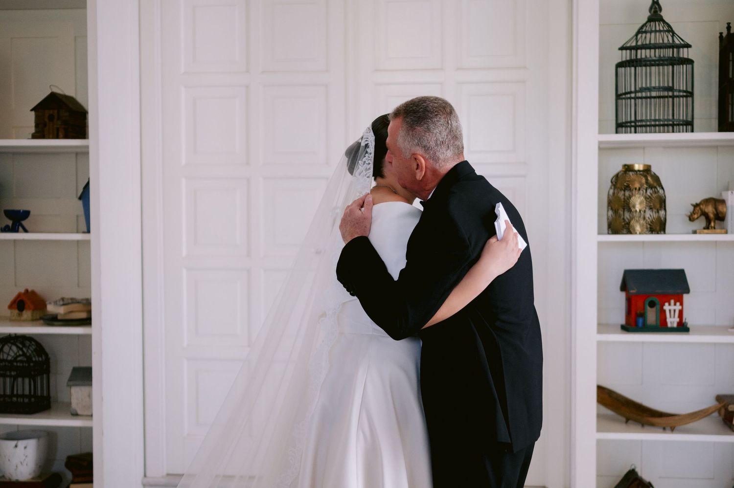 An emotional embrace during a wedding moment in front of white built-in shelves with decorative items.