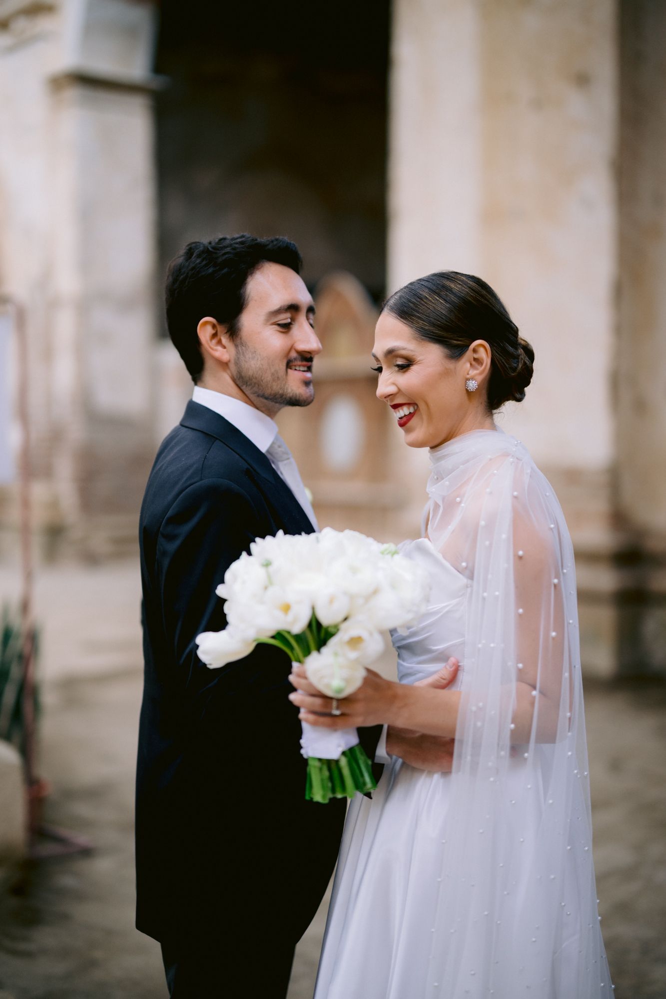 Intimate moment between newlyweds sharing laughter while holding white bouquet.