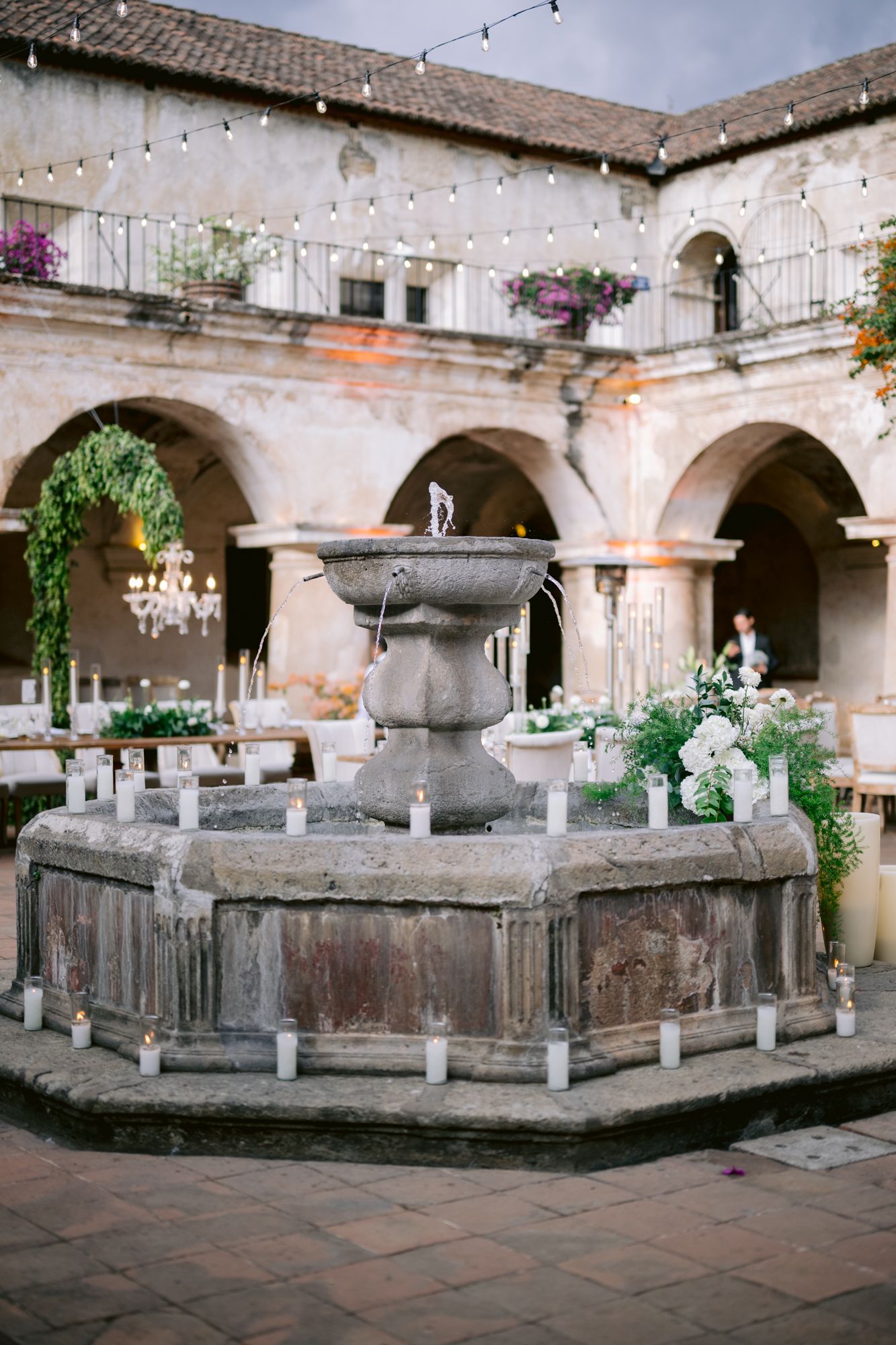 Historic stone fountain centerpiece in courtyard surrounded by arched columns and hanging plants.