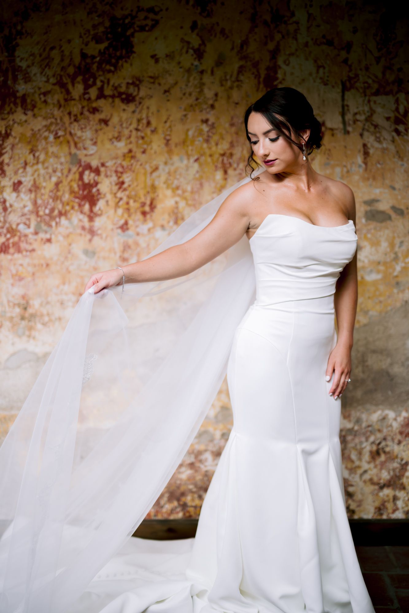 An elegant bride in a fitted white gown arranges her flowing veil against a weathered wall.