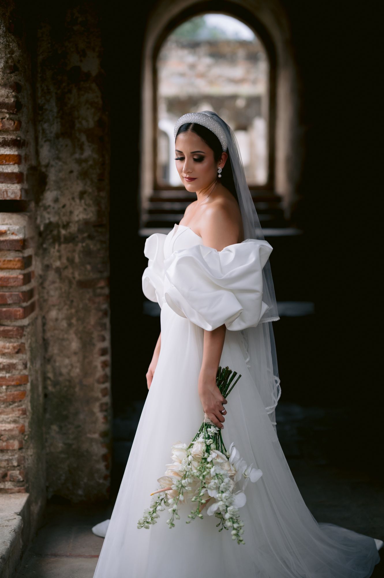 A bride in an elegant off-shoulder gown poses in an arched doorway holding white flowers.
