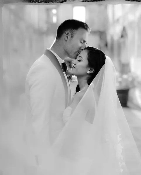 A tender romantic moment between newlyweds as they embrace, with the bride wearing a flowing white veil in black and white.