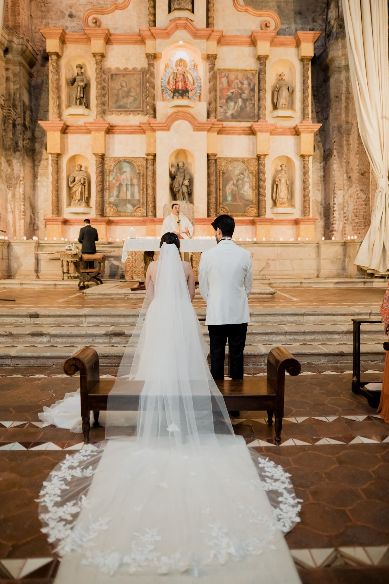 Long cathedral veil trailing behind couple as they face ornate religious altar in historic church.