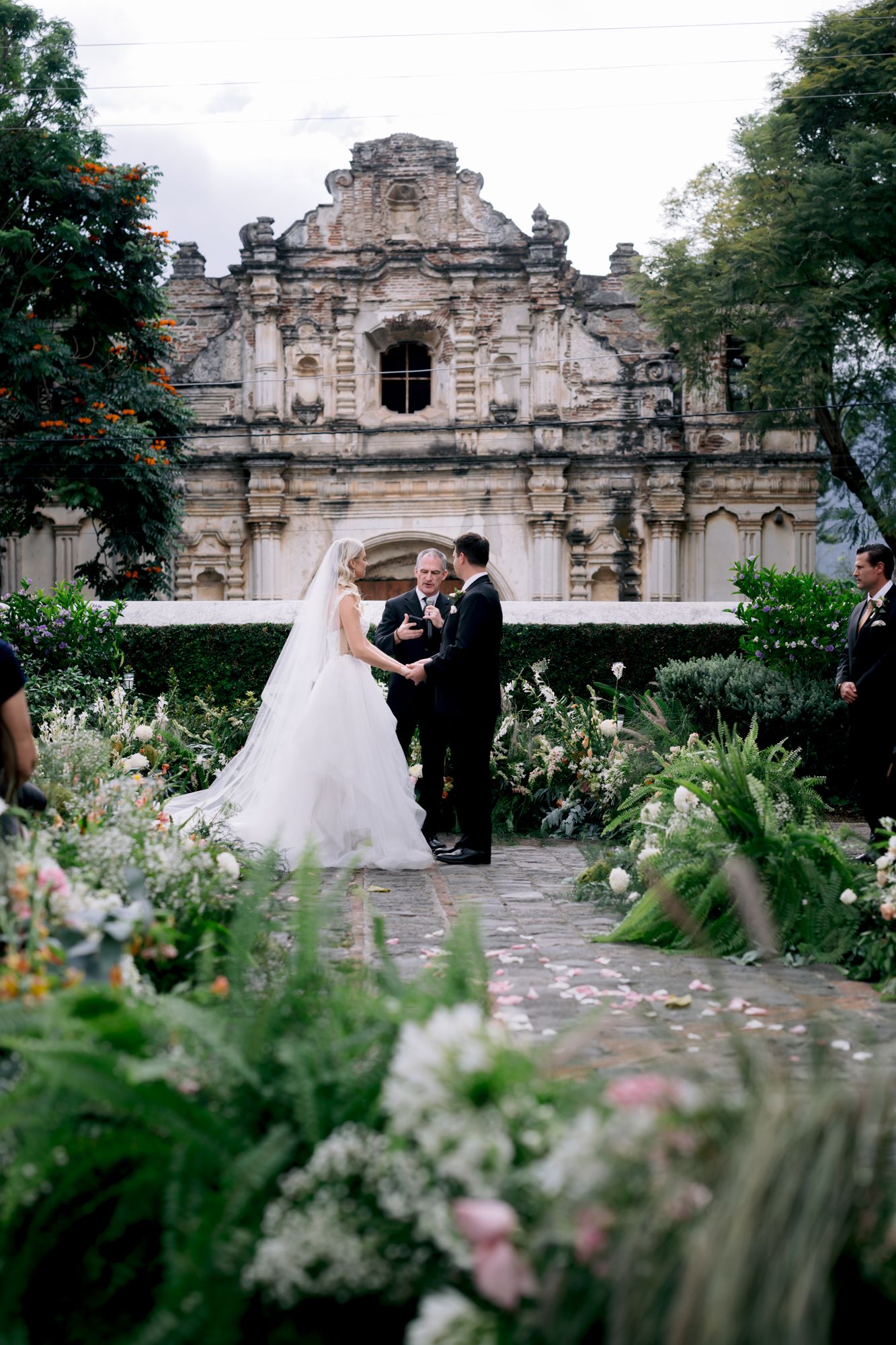 Wedding ceremony with dramatic stone church backdrop surrounded by flowering garden paths.
