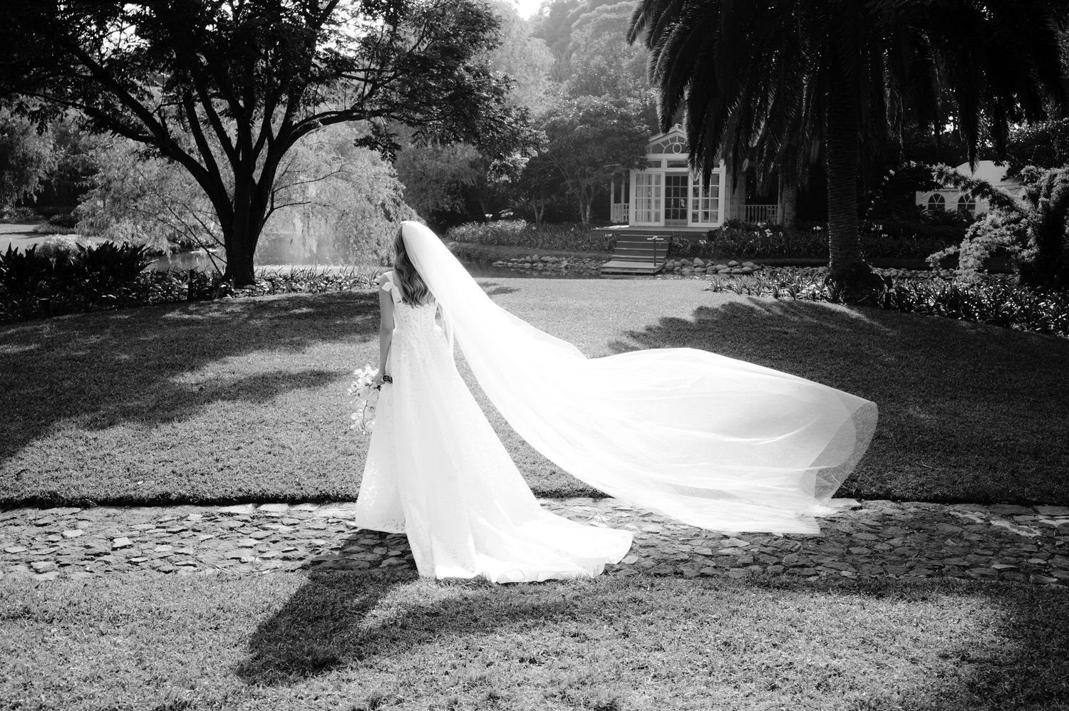 A flowing white wedding dress train trails across a grassy lawn in black and white photography.