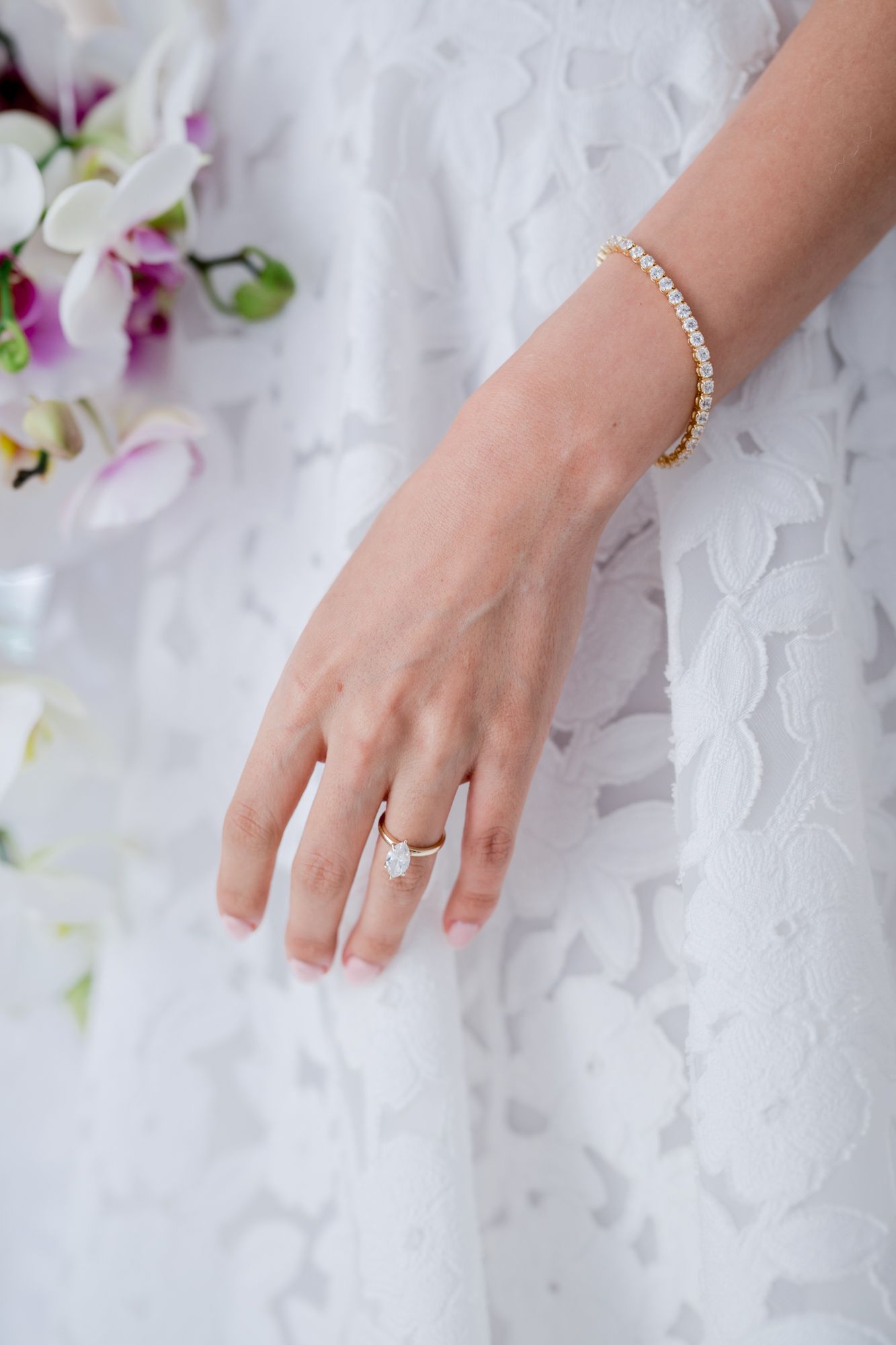 Close-up of hands wearing wedding rings and bracelet against white lace dress fabric with orchid flowers.