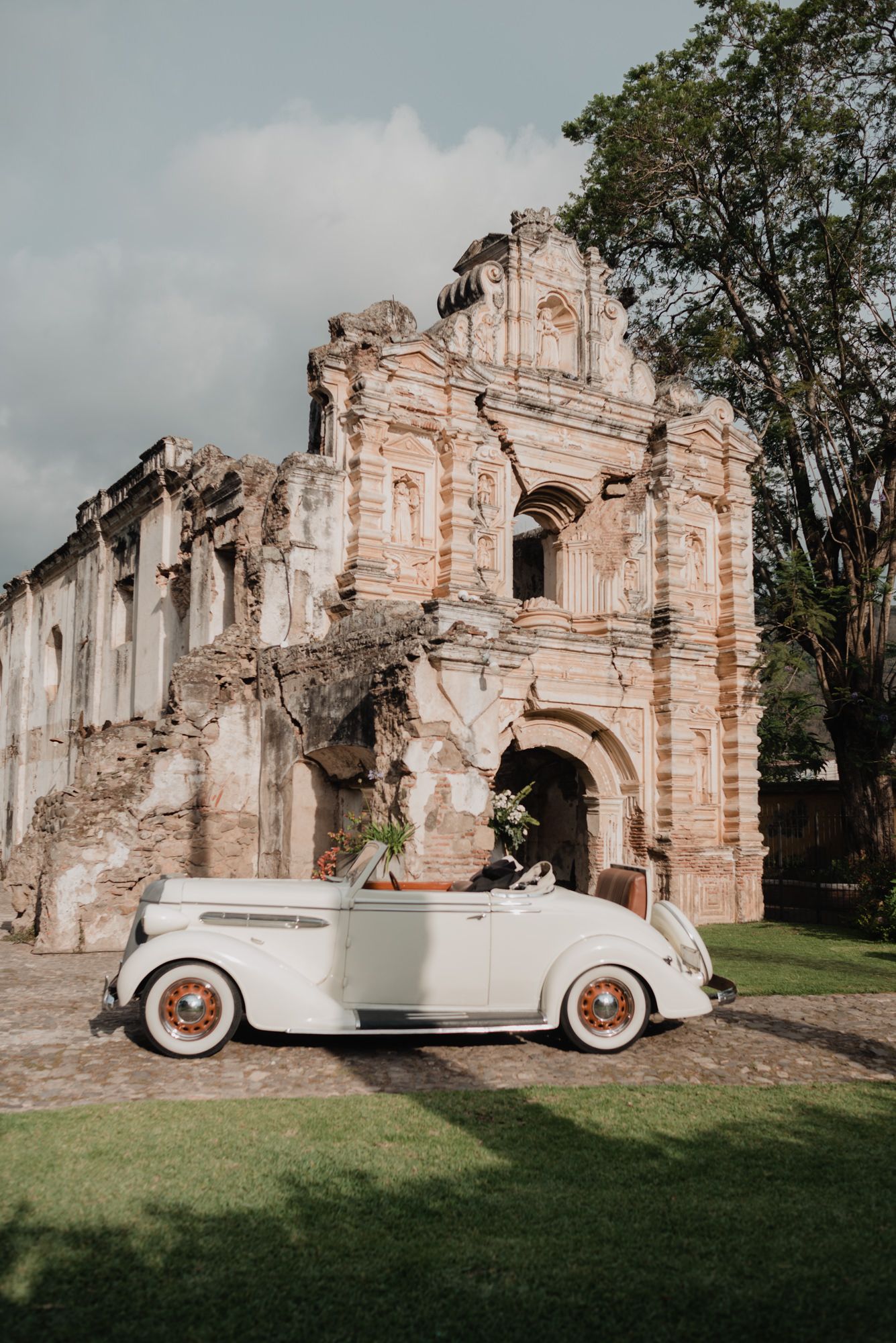 Vintage white luxury car parked in front of historic stone ruins and archways on grassy grounds.