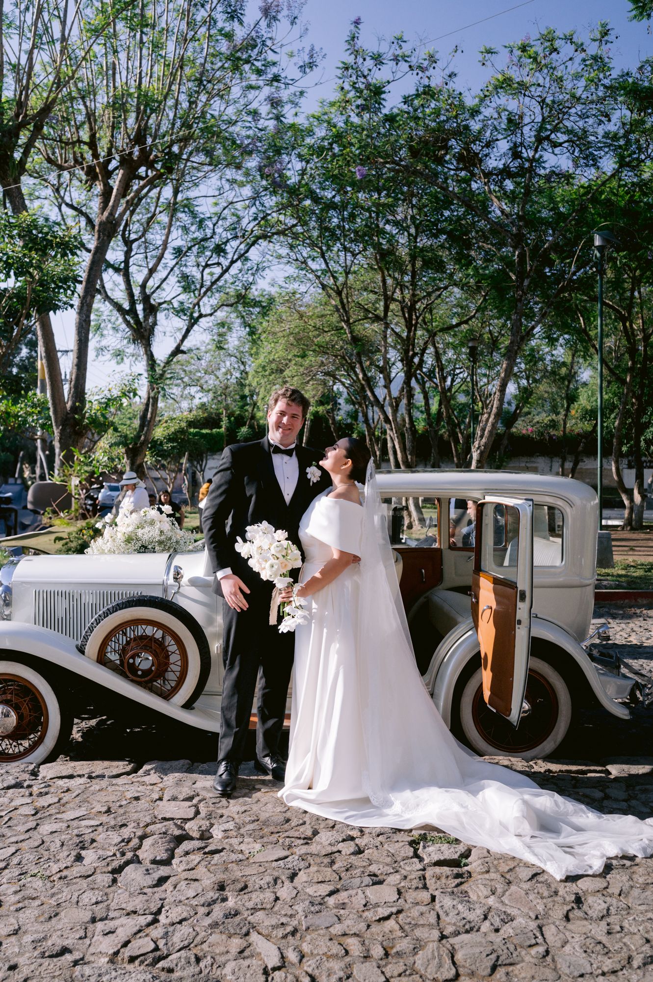 A classic vintage car serves as backdrop for a wedding couple posing on cobblestone pavement surrounded by trees.
