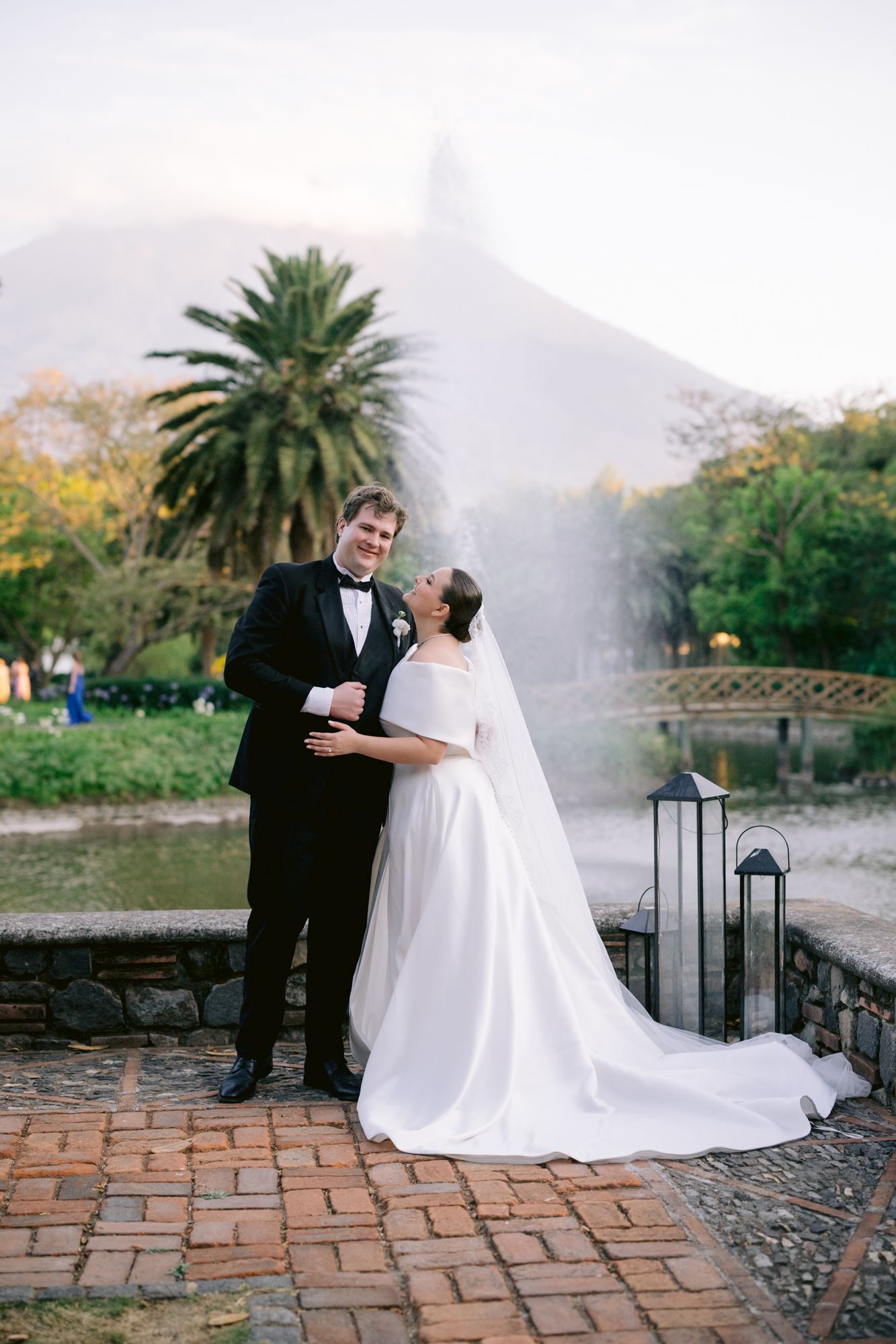 Newlyweds share romantic moment by fountain with mountain silhouette and palm trees in background.