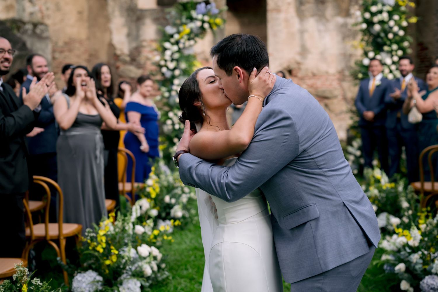 Happy couple shares their first kiss as guests cheer during an outdoor wedding ceremony with floral decor.