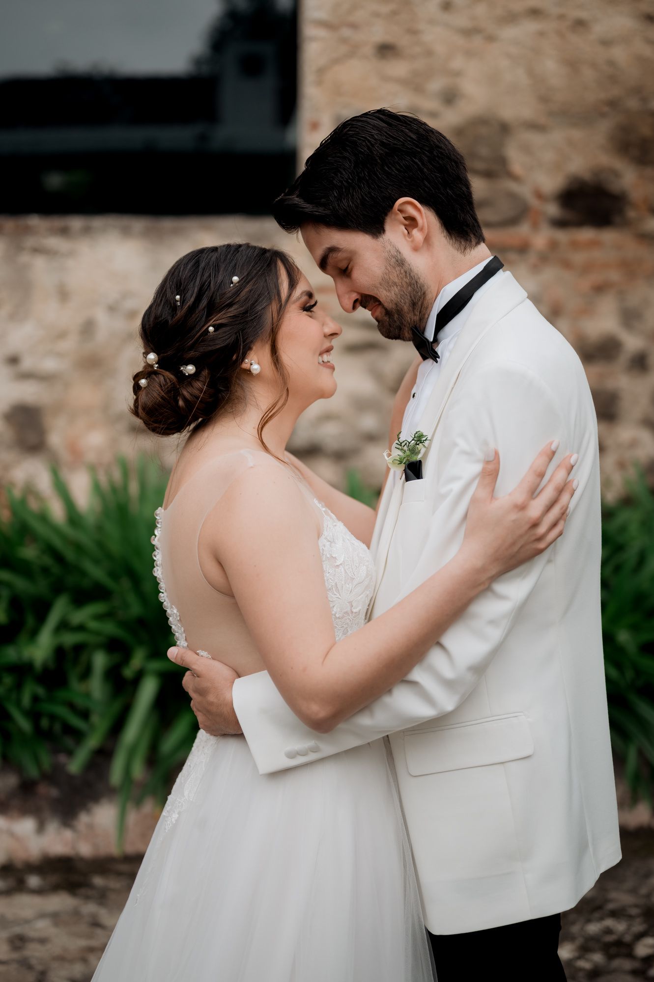 Wedding couple shares romantic embrace against rustic stone wall and garden setting.