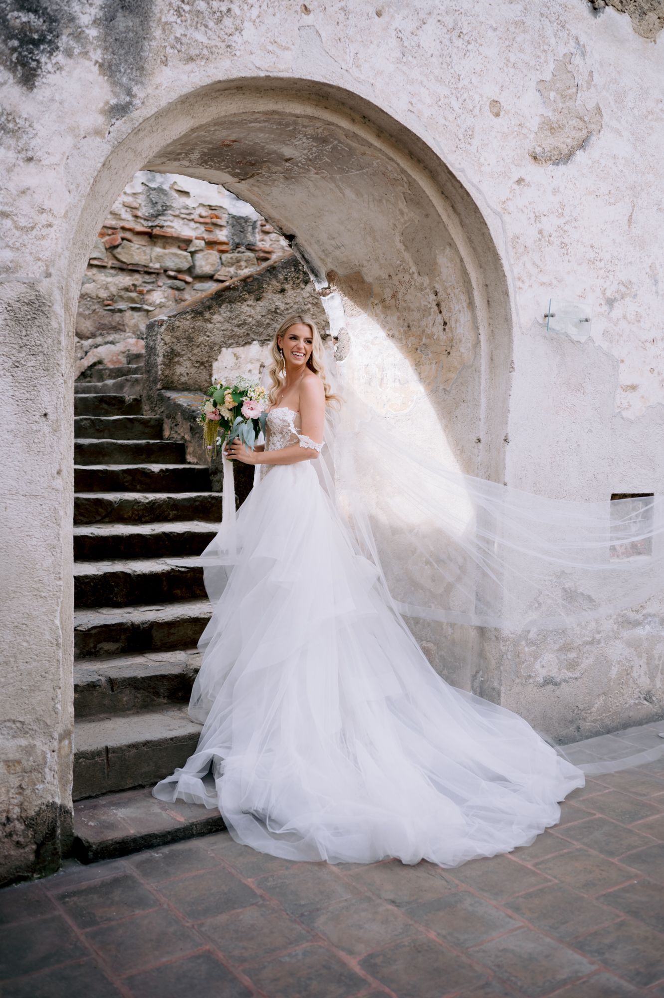 Dramatic full-length bridal portrait under stone archway with cascading train and bouquet.
