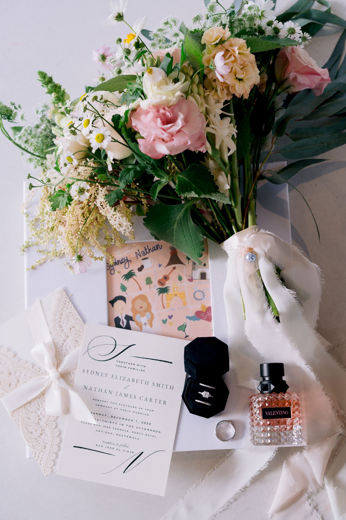 Wedding details flatlay with flowers, invitation, perfume bottle and rings.