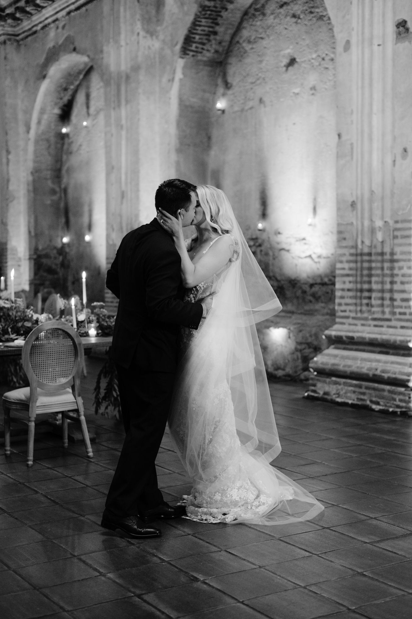 A romantic black and white wedding photo shows a couple sharing a kiss in a historic venue with candles.