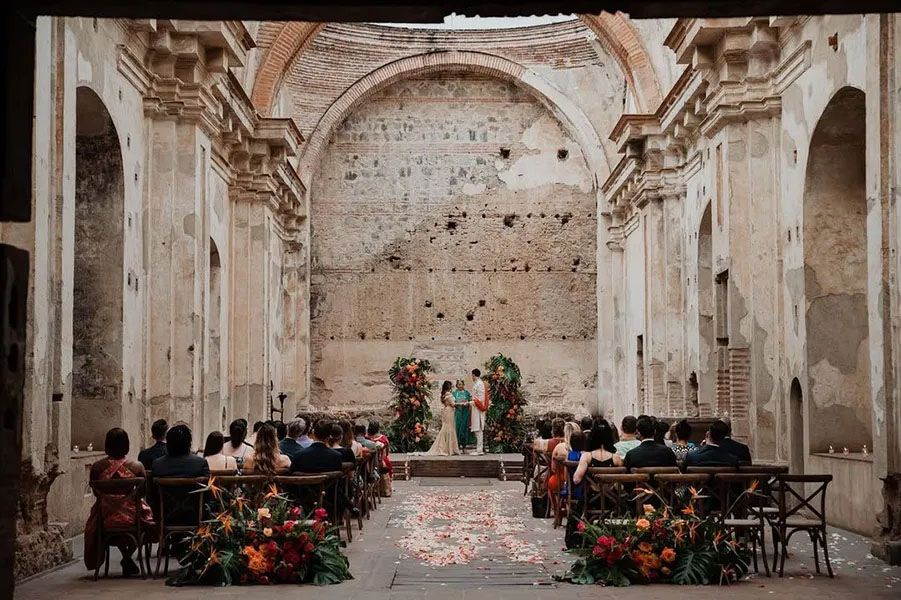 Intimate wedding ceremony in a rustic stone chapel with exposed walls and floral decorations lining the aisle.