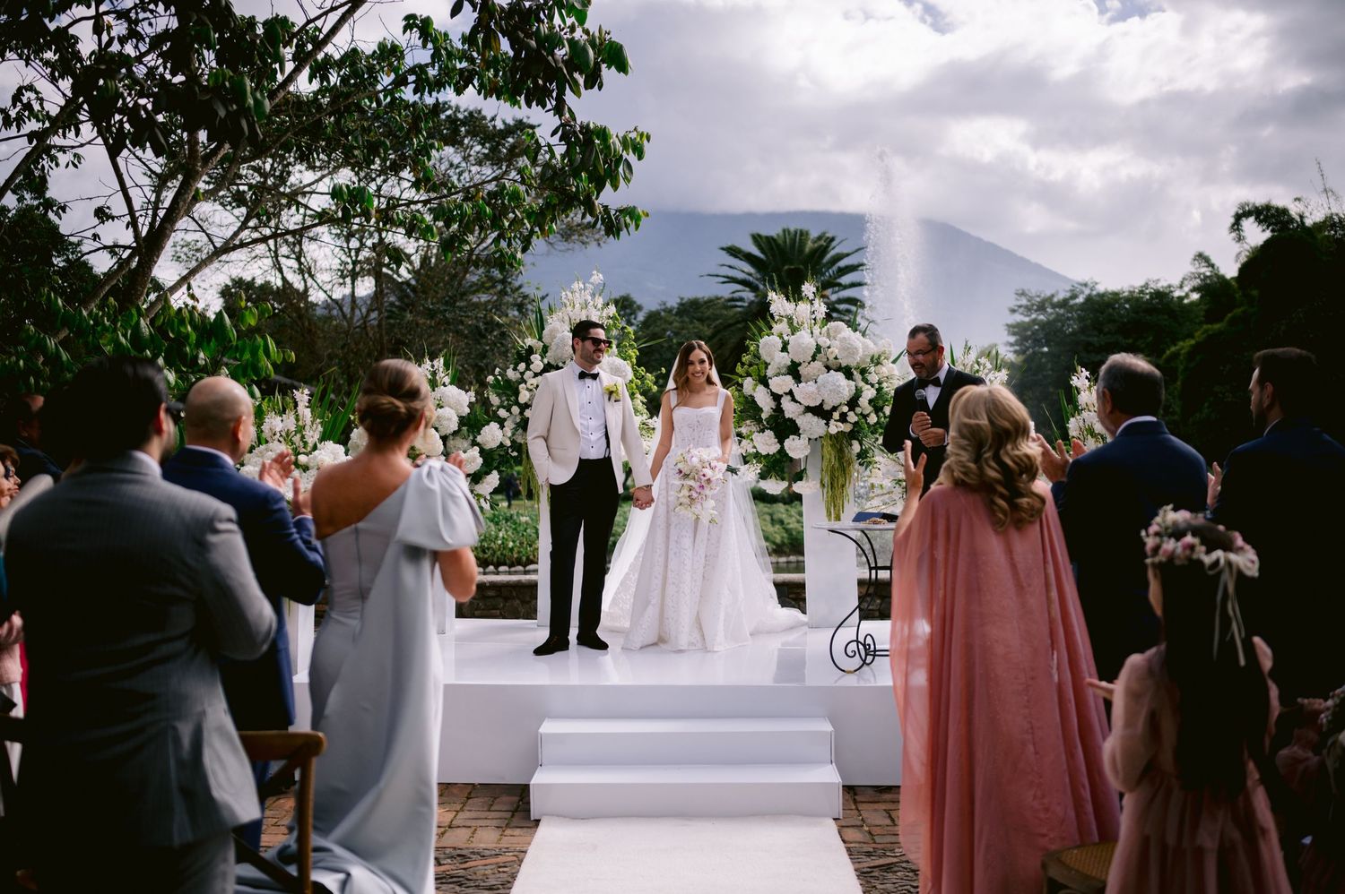 Outdoor wedding ceremony setup with white columns and floral arrangements under cloudy skies.