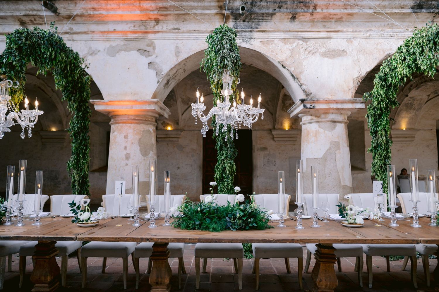 Long rustic dining tables set under crystal chandeliers and ivy-covered arches.