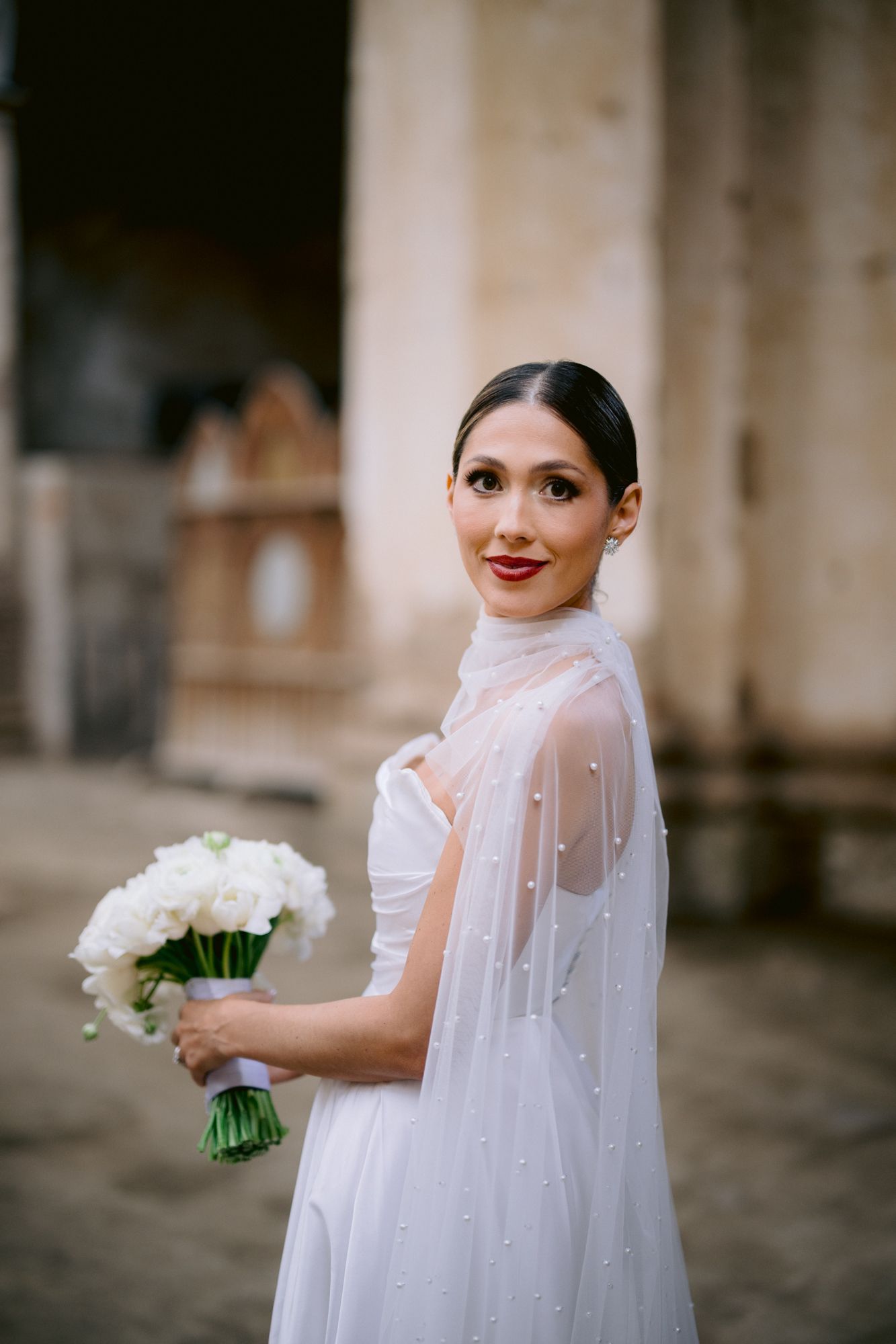 Bride in modern white gown and pearl cape holding bouquet while smiling at camera.
