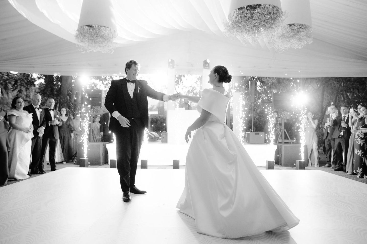 A black and white wedding photo captures a romantic first dance moment under dramatic ceiling lighting.