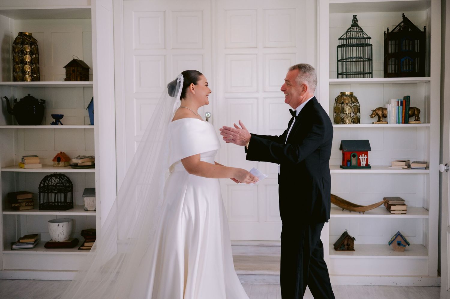 A tender pre-wedding moment between a bride and gentleman in front of decorative shelving with birdcages.