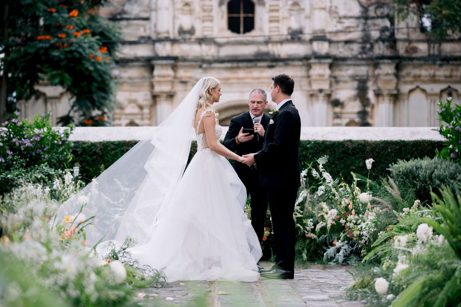 Wedding ceremony in progress at rustic stone church surrounded by lush garden landscaping.