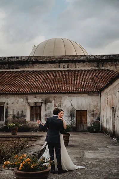 A couple in wedding attire embraces in front of a weathered building with a domed roof and terra cotta tiles.