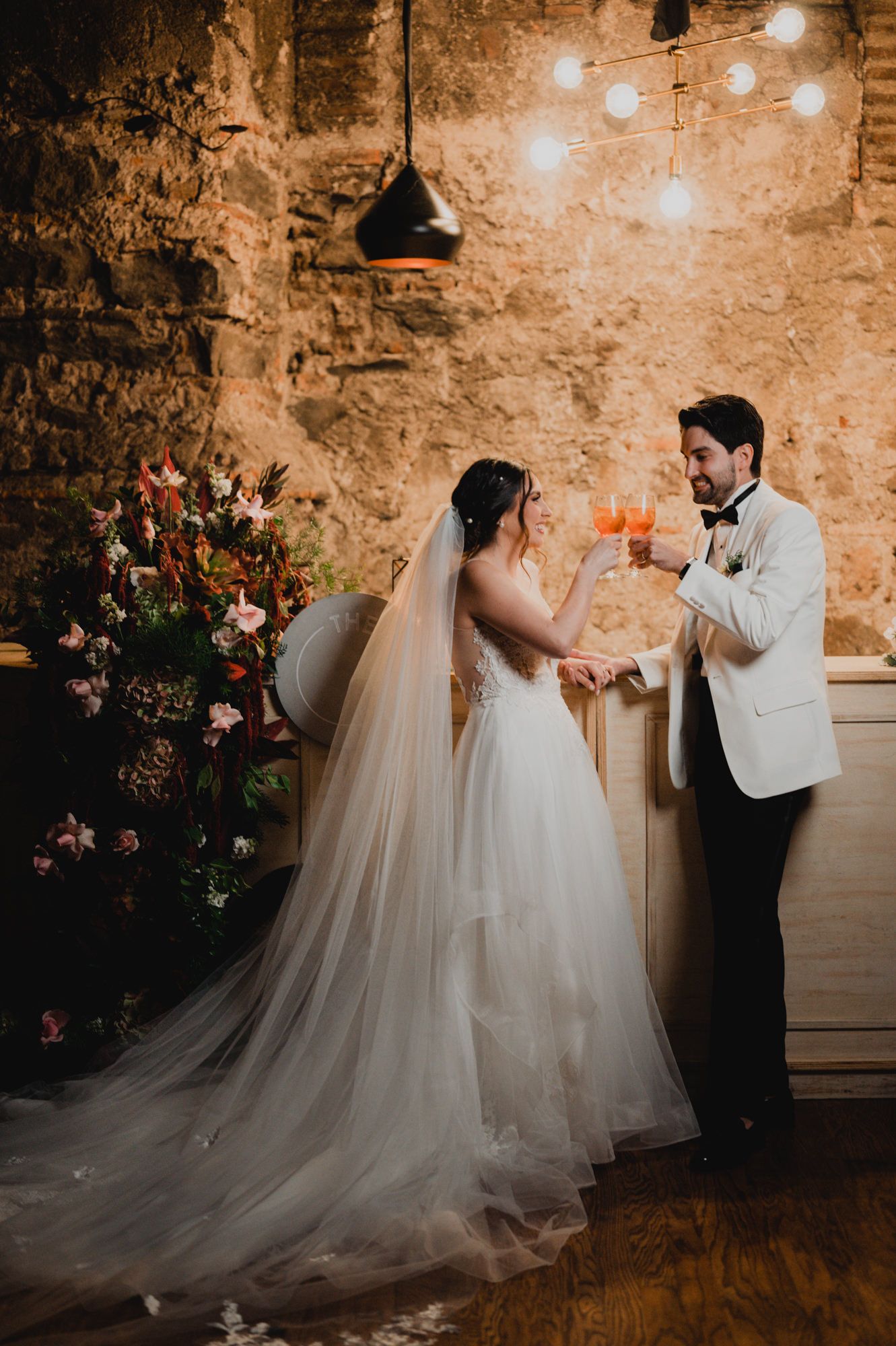 Newlyweds toast with drinks against rustic stone wall backdrop.