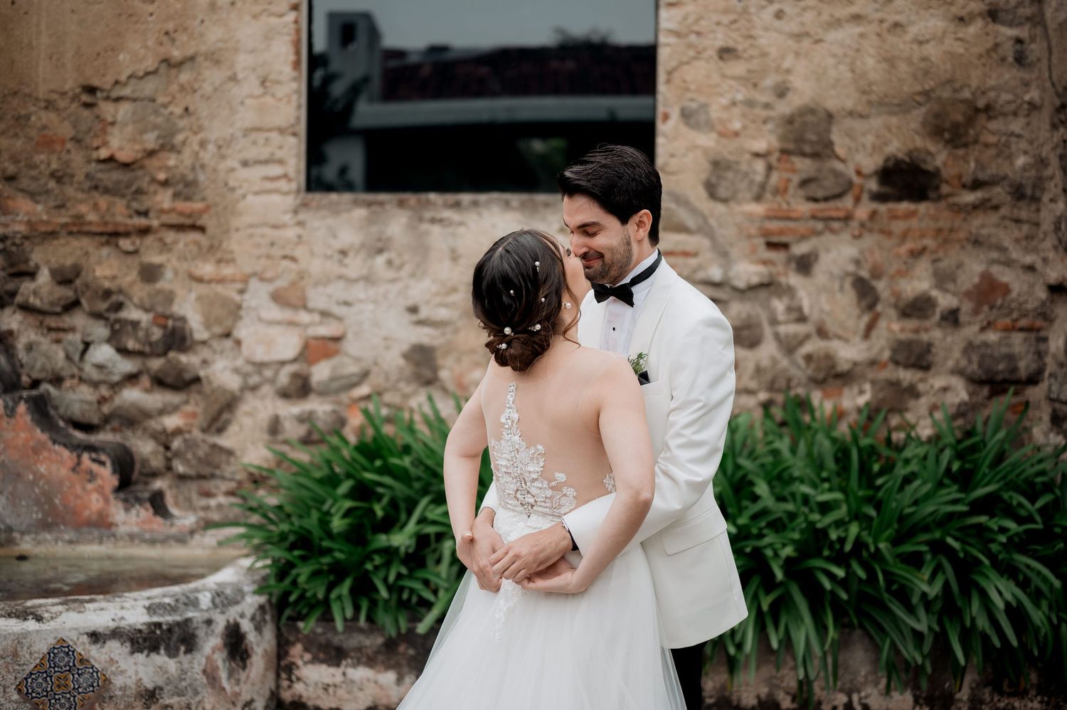 Intimate wedding couple embrace in front of stone wall with garden backdrop.