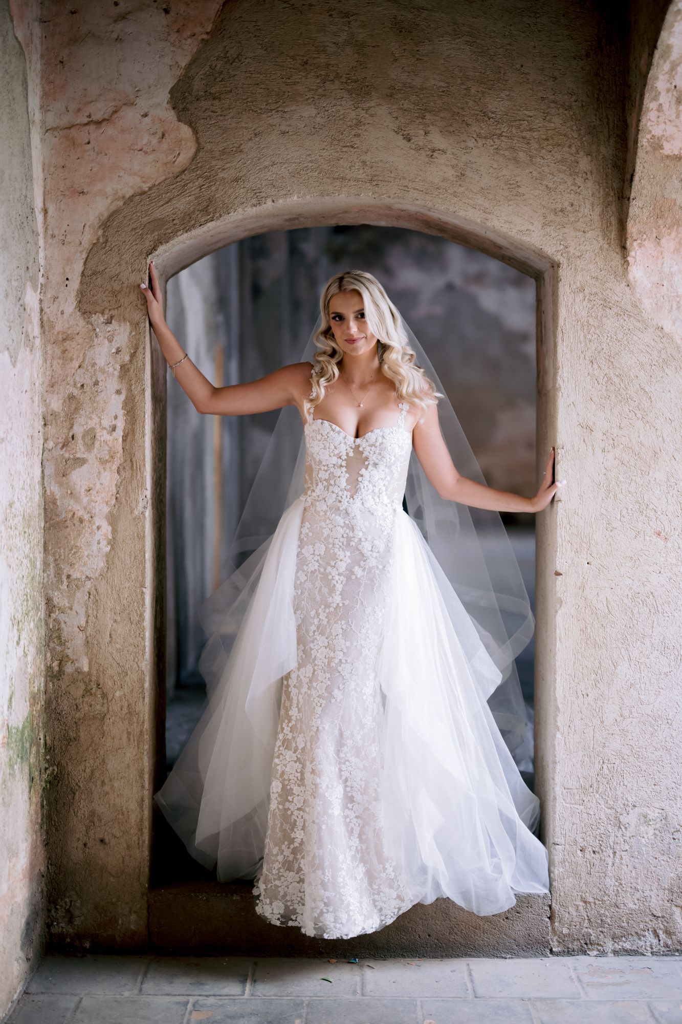 A bride in a lace wedding gown poses elegantly in a stone archway.