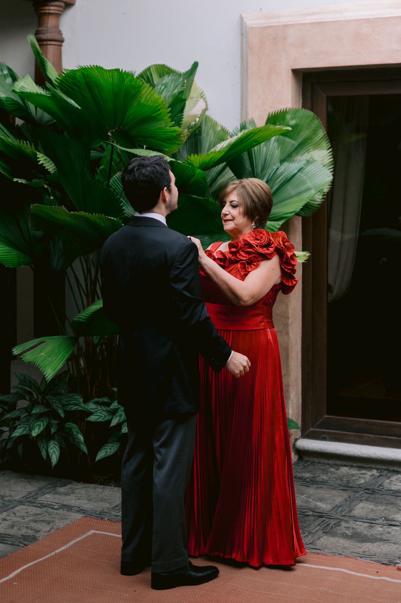 A couple in formal attire stand together against lush green tropical plants.