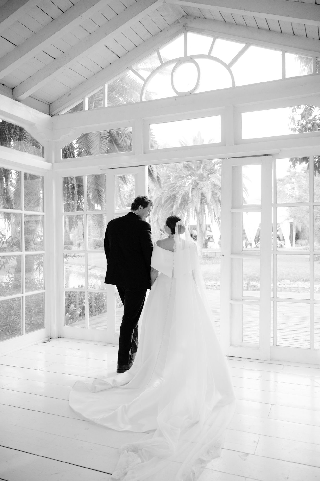 Black and white photo of newlyweds standing together in front of dramatic floor-to-ceiling windows.