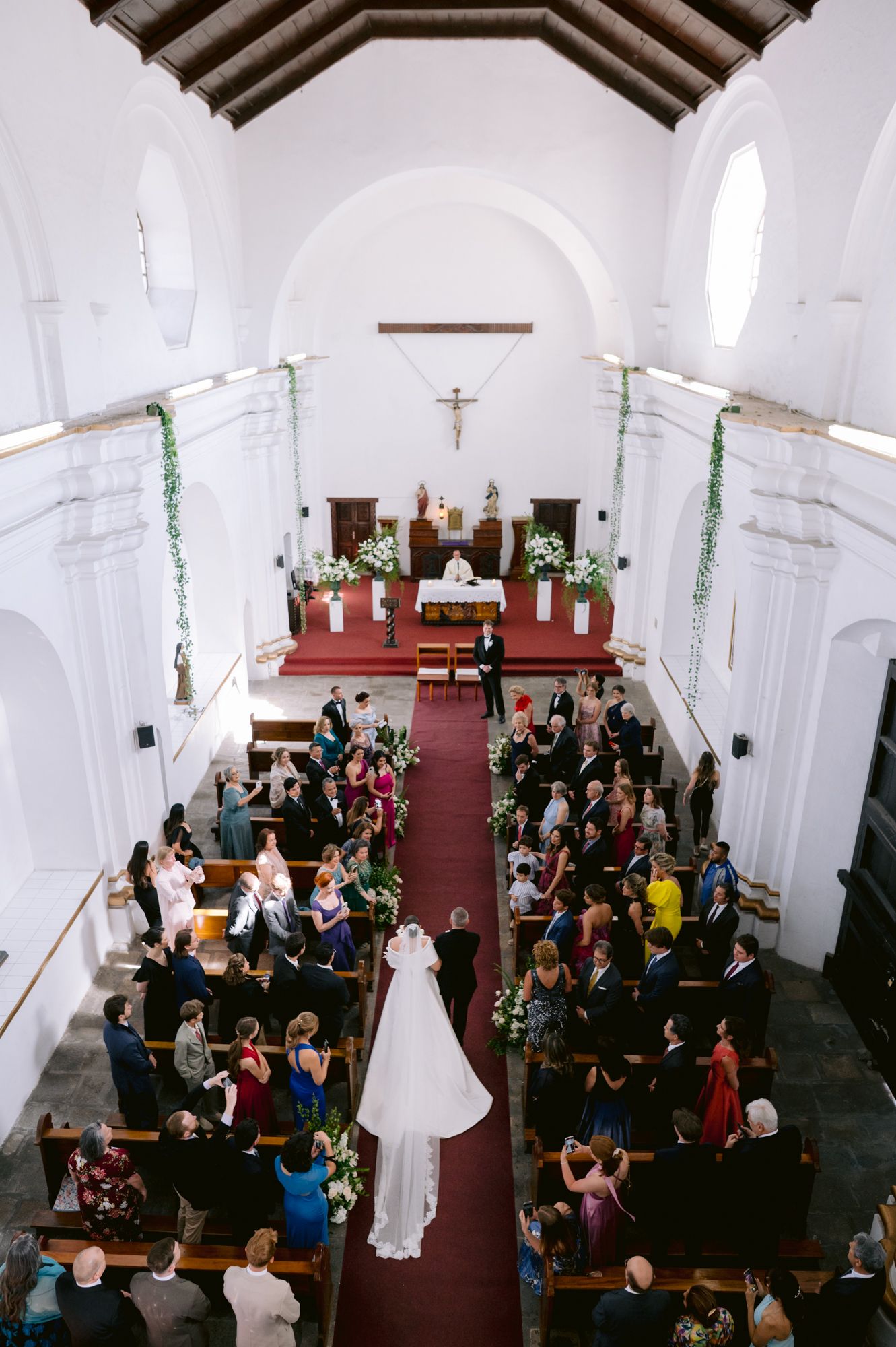 Aerial view of a wedding ceremony in a traditional white church with red carpet and wooden pews.