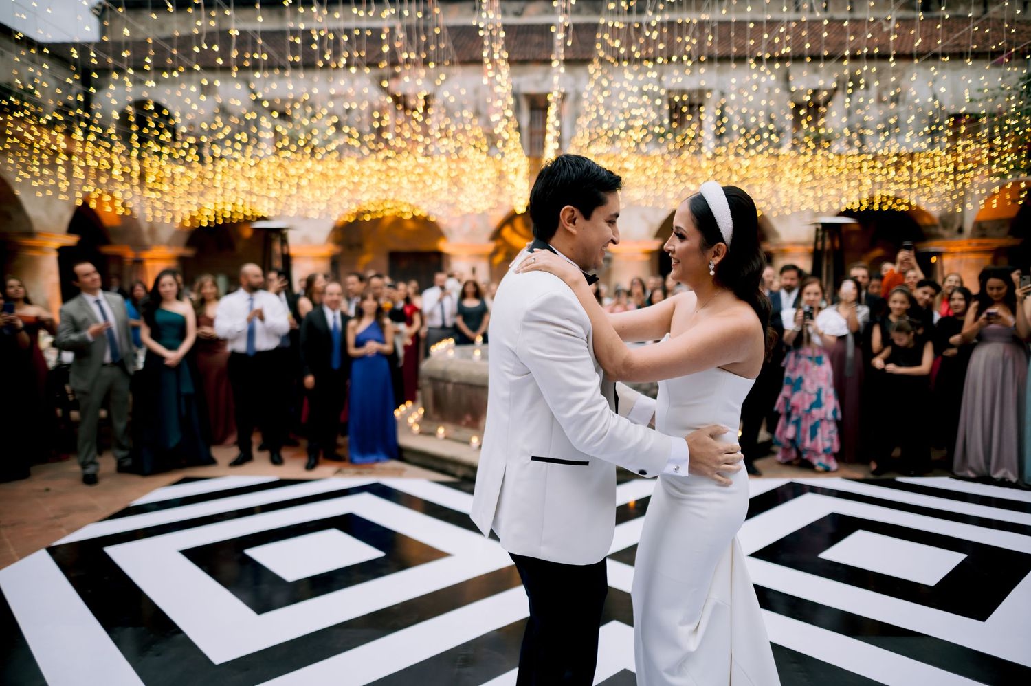 Newlyweds share first dance on striking black and white patterned floor under twinkling lights.