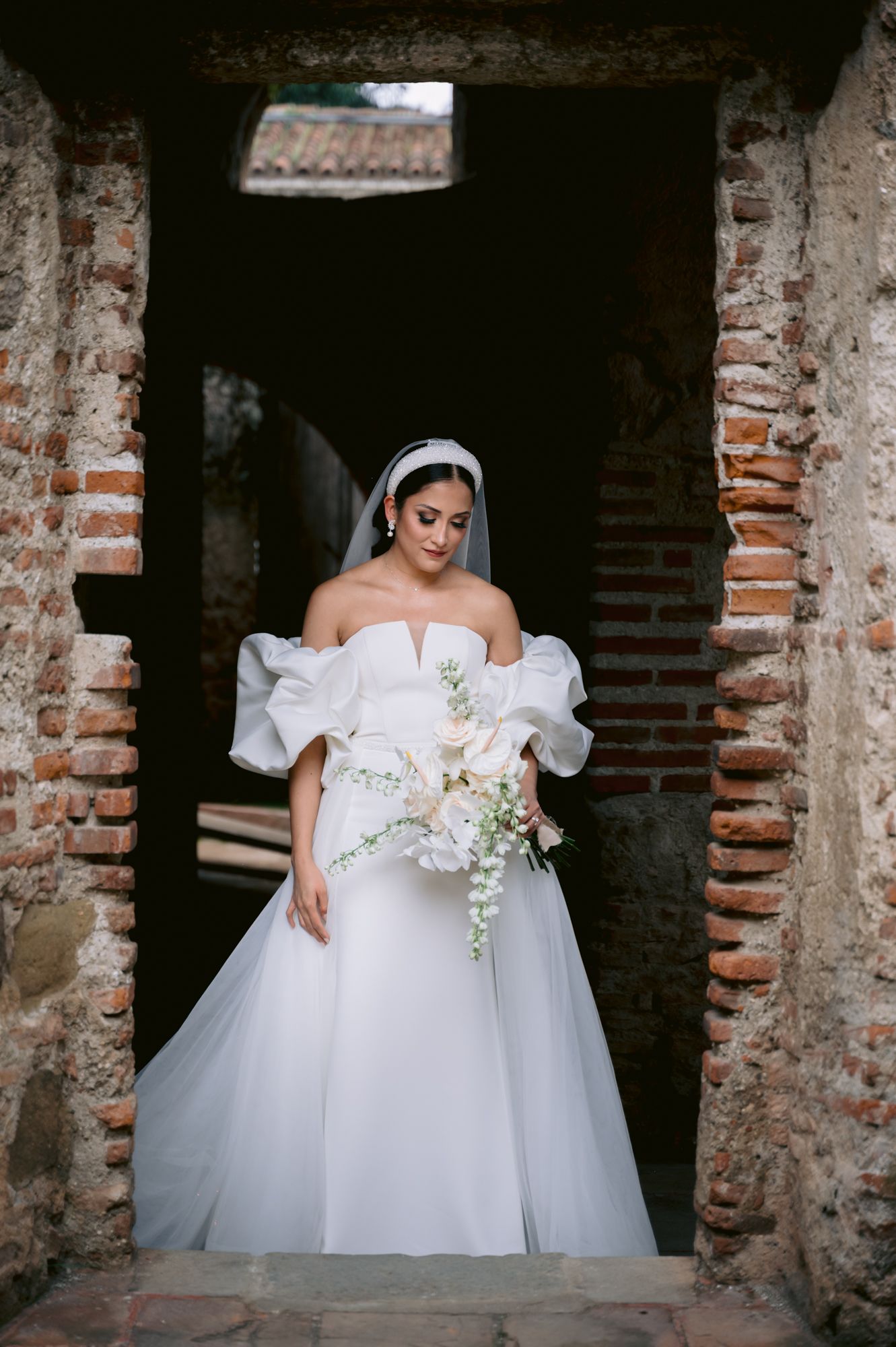 Elegant bride in off-shoulder white gown with puff sleeves stands in rustic brick doorway holding cascading bouquet.