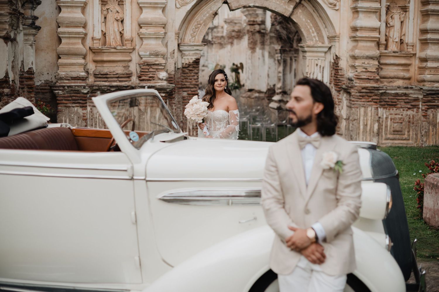 Classic white vintage car parked near stone ruins creates a romantic wedding scene.