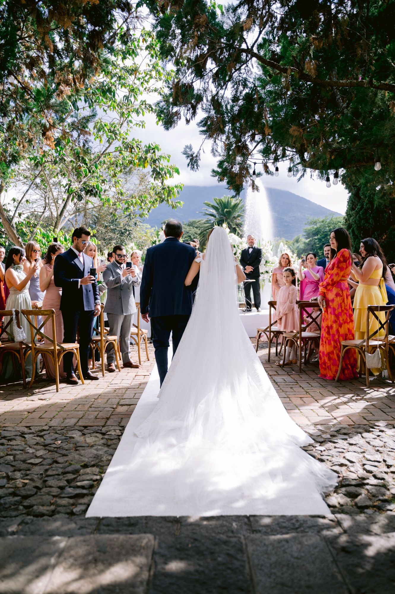 Wedding ceremony in progress on a stone paved courtyard with guests seated on wooden chairs and mountain views.