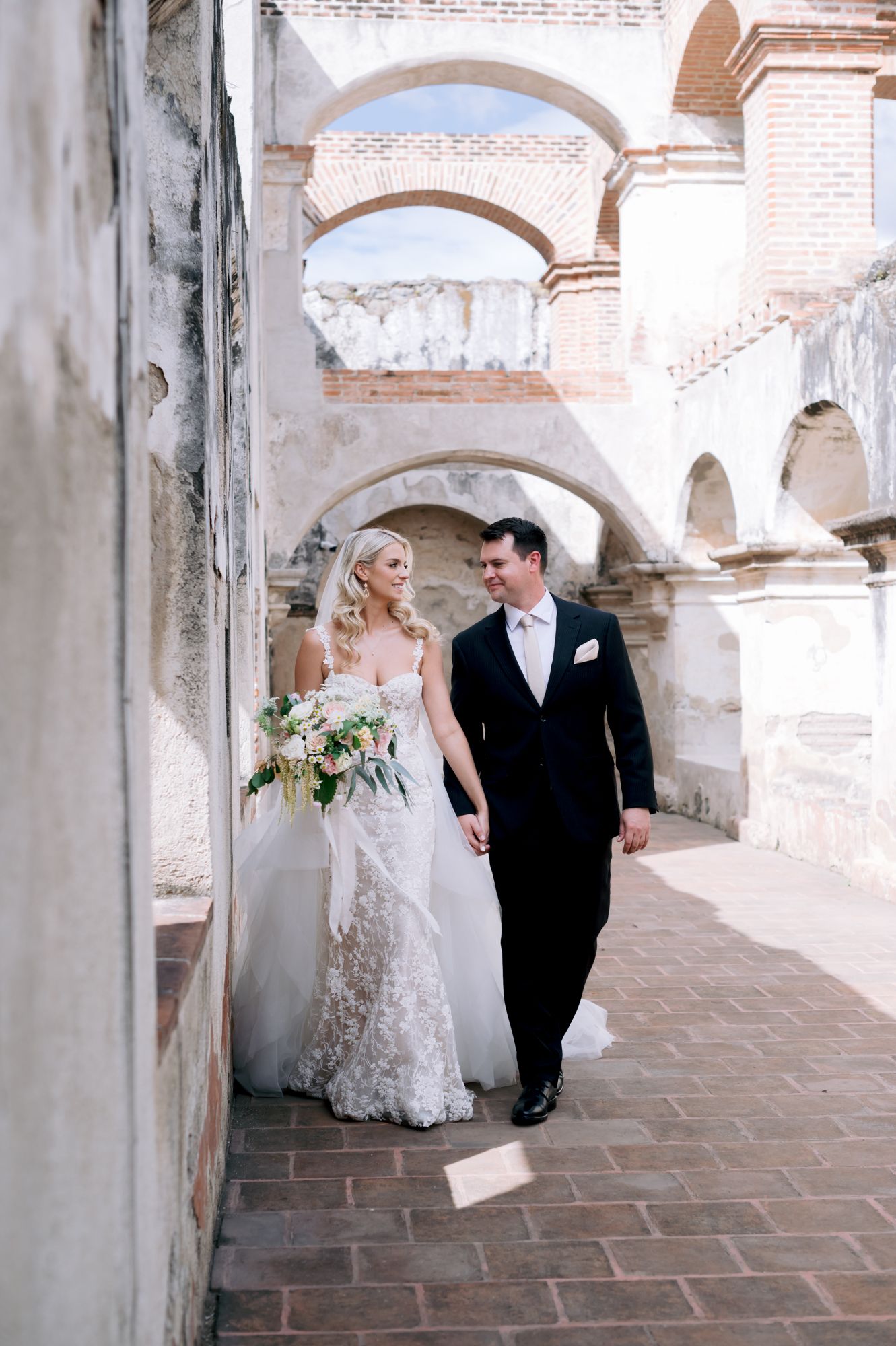 Wedding couple strolling beneath rustic stone arches in a romantic European setting.