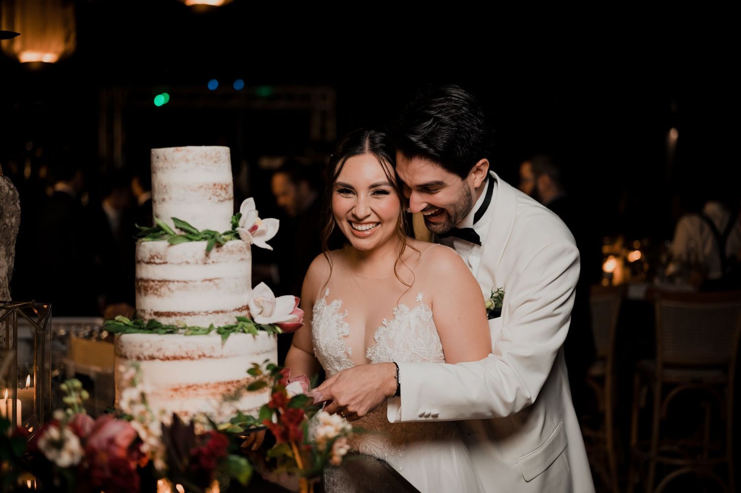 Wedding couple posing next to their elegant three-tiered white wedding cake with floral decorations.