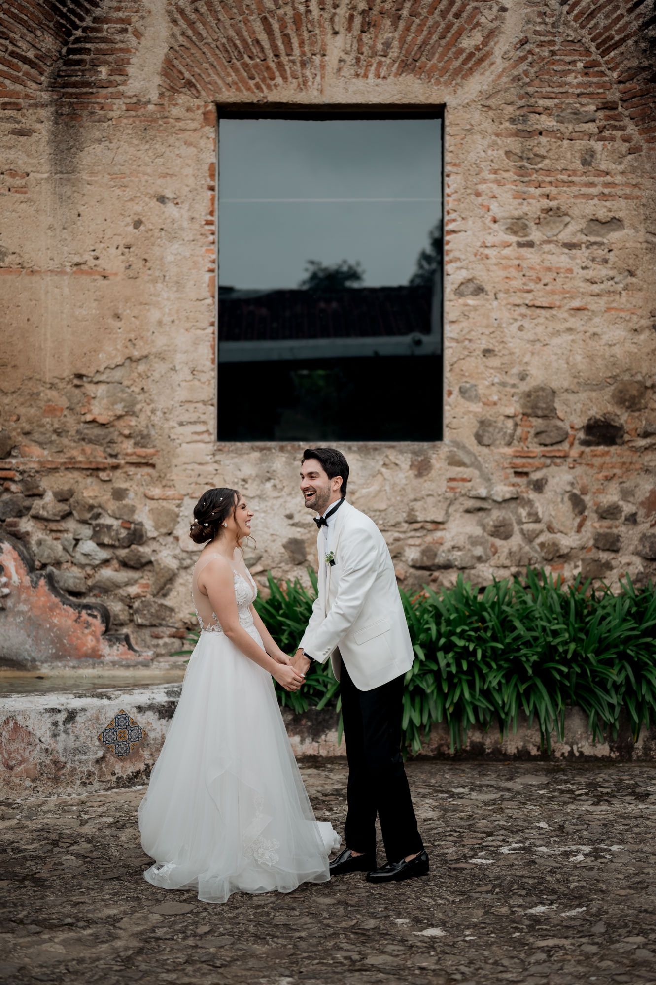 Wedding couple laughs together in front of historic stone architecture and greenery.