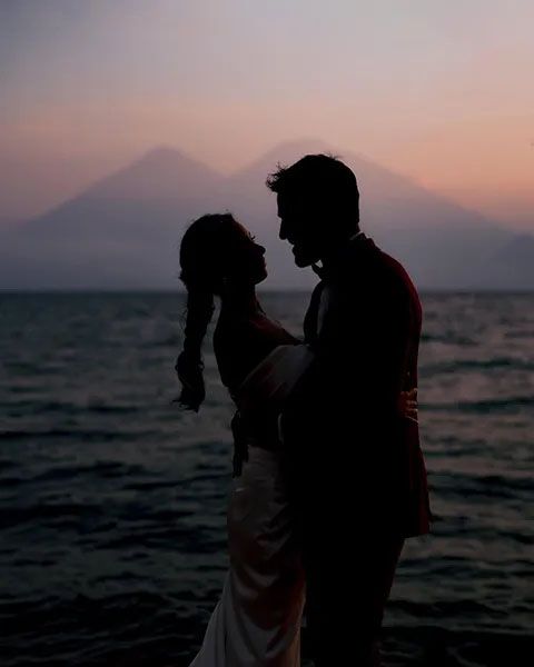 Romantic silhouettes stand against a dreamy sunset backdrop with mountains and ocean waves at dusk.