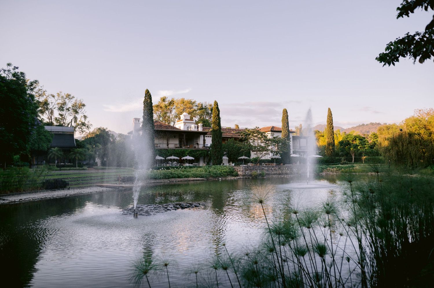 Serene lake view with fountains and elegant building at sunset surrounded by lush landscaping.
