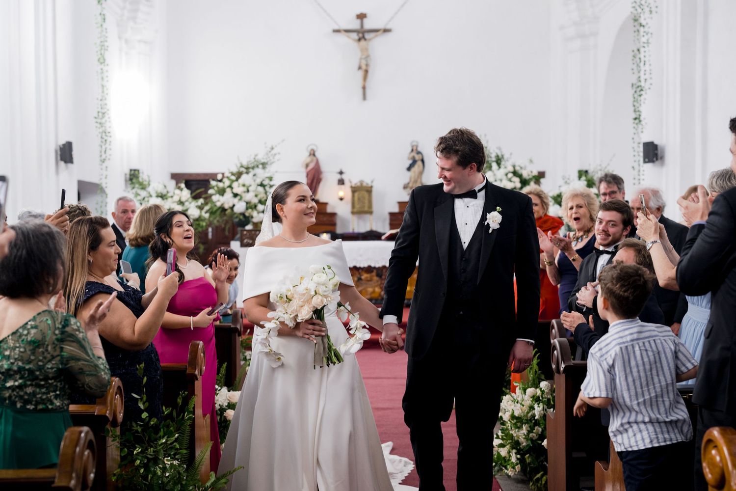 Newlyweds walk down the church aisle surrounded by applauding guests and beautiful floral arrangements.
