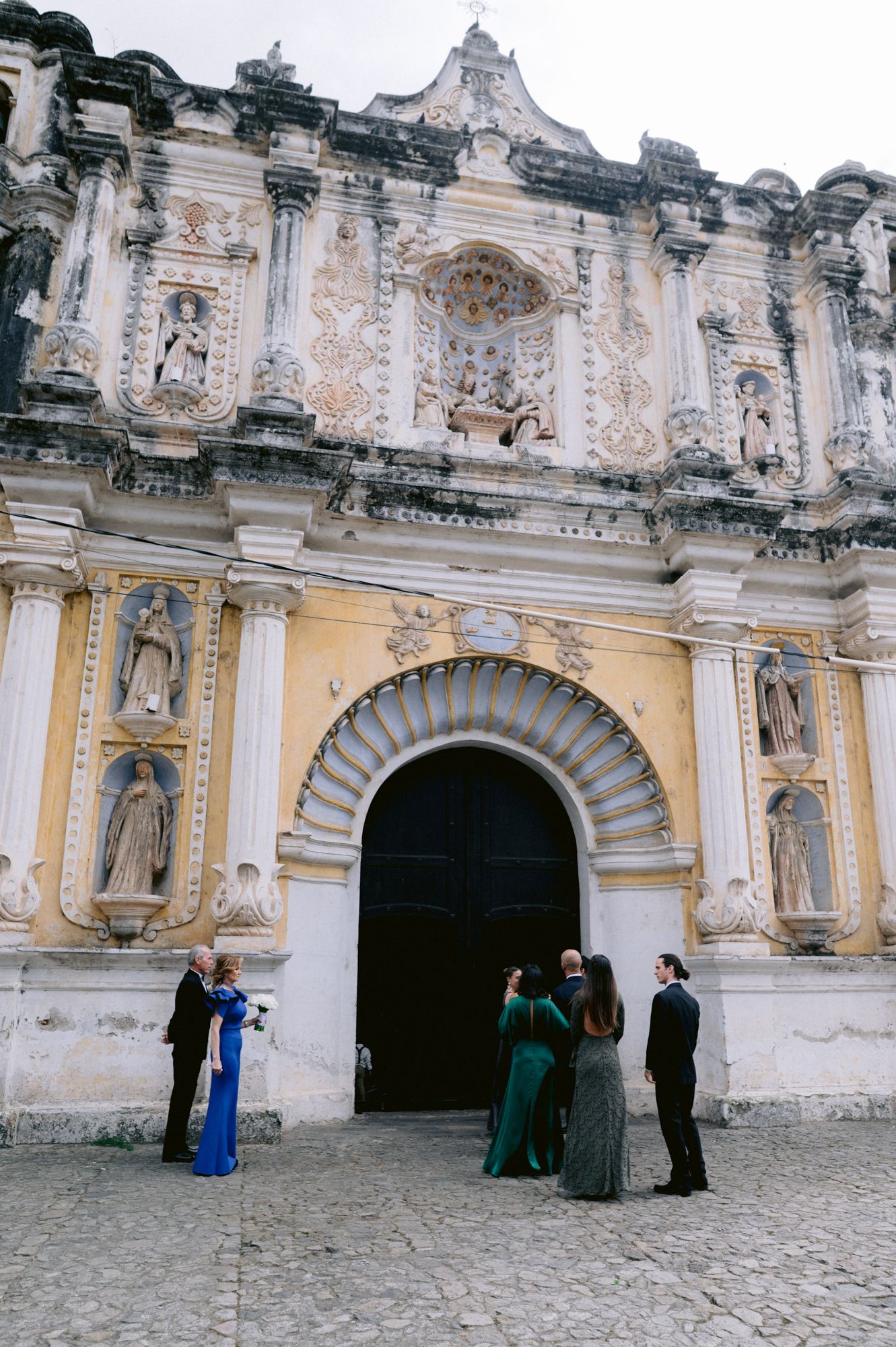 Wedding guests gathering outside an ornate colonial church facade with baroque architectural details.
