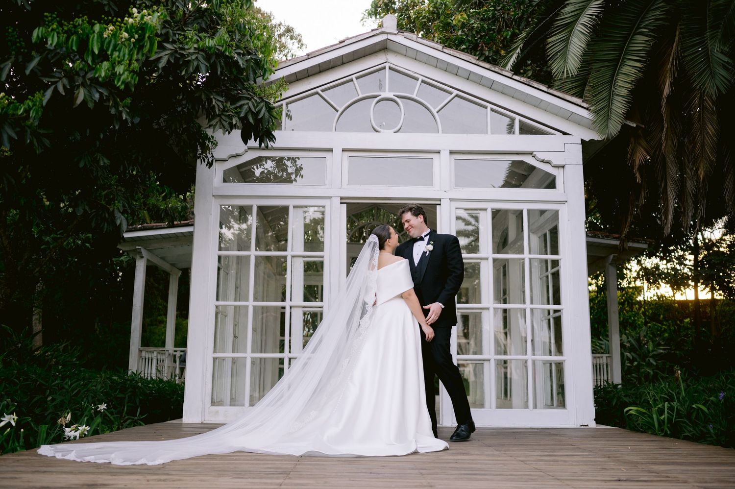 Wedding couple poses in front of a white glass conservatory surrounded by lush tropical gardens.