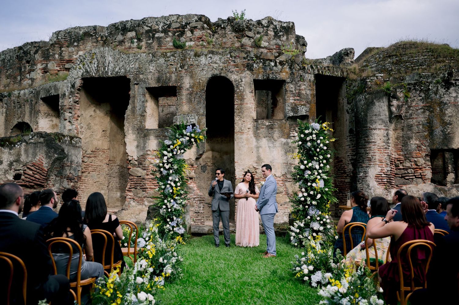 Wedding ceremony setup with floral arches in historic ruins with guests seated on wooden chairs.